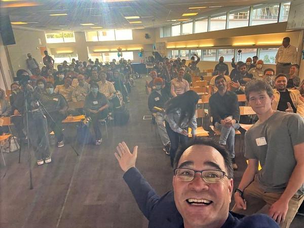 A dark haired man dressed in a sport coat and tshirt, laughing, takes a selfie with an amber-toned roomful of people behind him. 